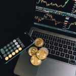 A still life of Bitcoin coins, a smartphone calculator, and a trading chart on a laptop.